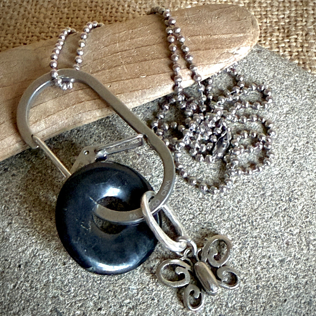 A silver-toned necklace with a large black shungite donut, a butterfly charm, and a carabiner-style clip, displayed on a textured gray surface with a wooden prop and burlap background.