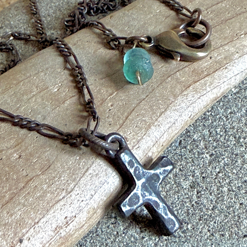 A close-up of a dark metal cross pendant necklace resting on a piece of weathered wood, with a small faceted teal bead and antique bronze clasp visible on the chain.