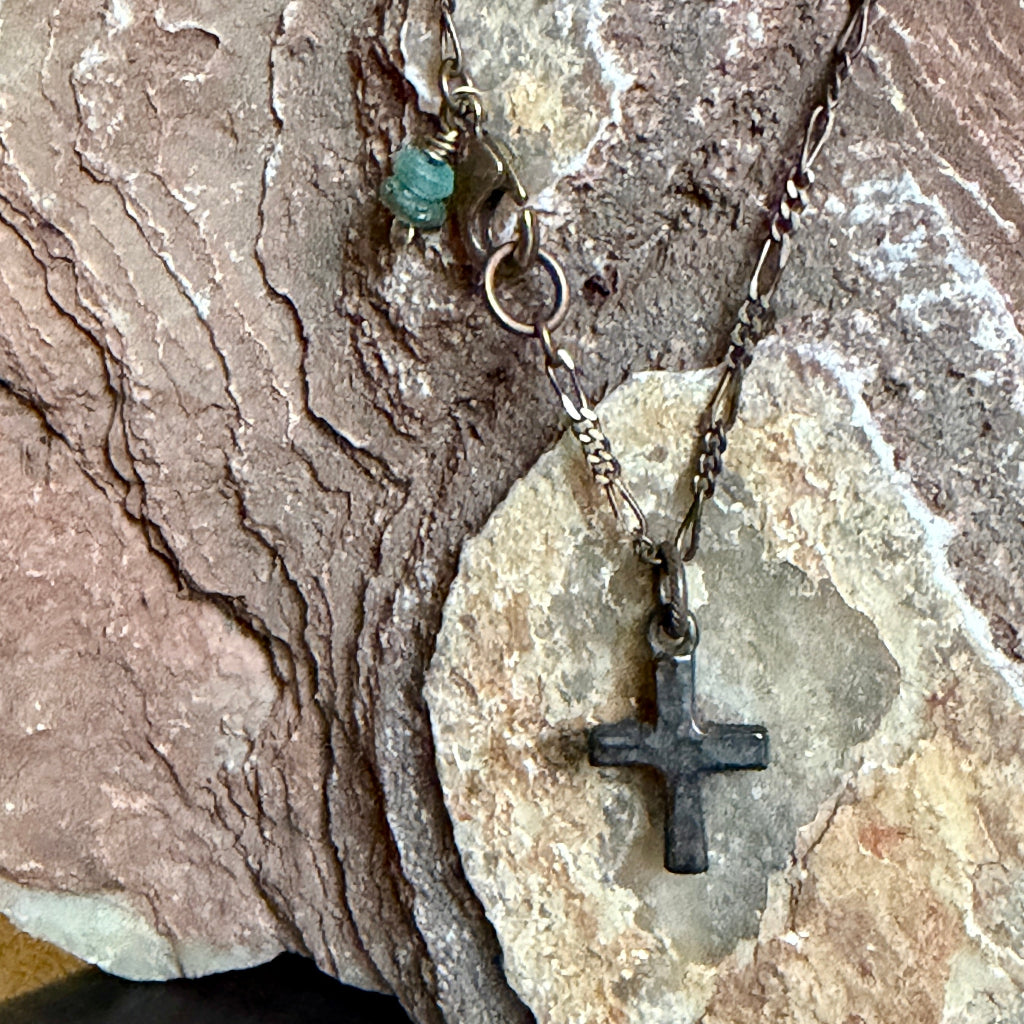 A dark cross pendant necklace with a textured metal chain and a small green bead charm, displayed on layered brown and beige rocks.