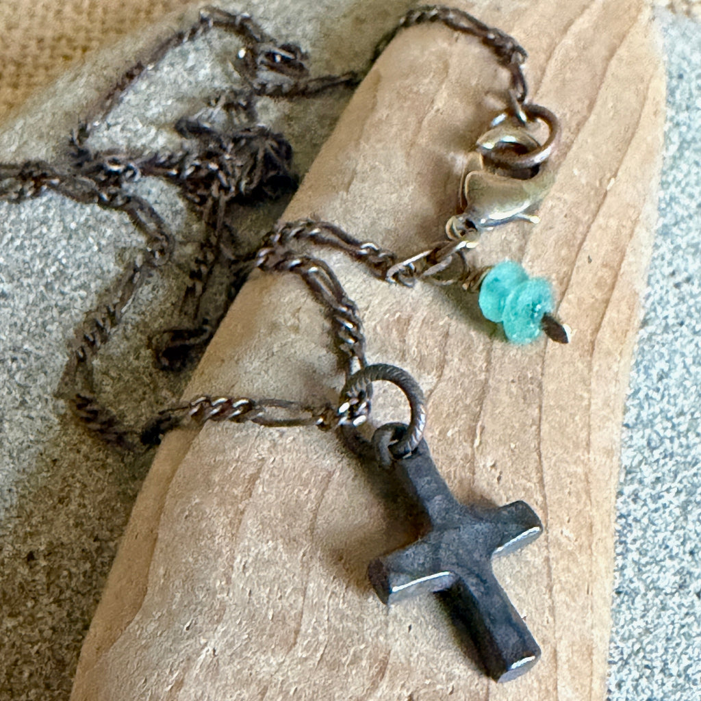 A dark metal cross pendant necklace with a figaro chain and a small turquoise glass bead dangle, displayed on a piece of wood.