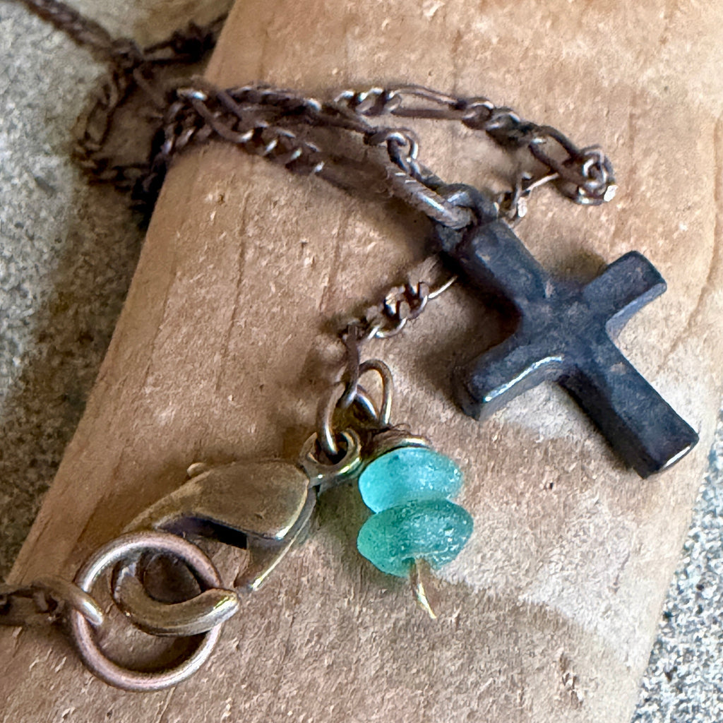 A close-up of a handmade necklace featuring a dark charcoal cross pendant, a bronze-toned lobster clasp, and two small translucent teal glass beads, displayed on a textured tan stone surface.