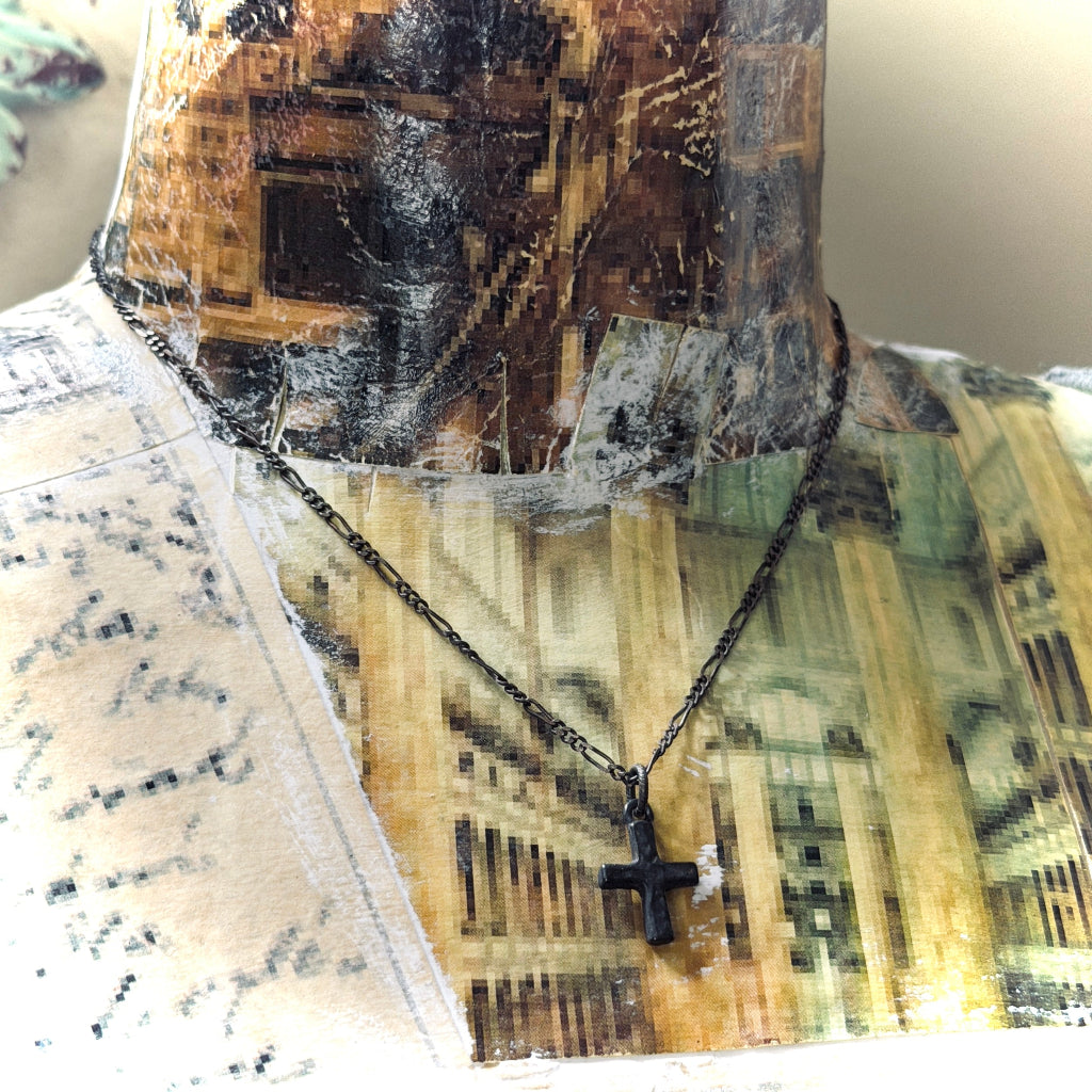 A black cross pendant necklace on a black chain, displayed on a vintage-style collage mannequin bust.