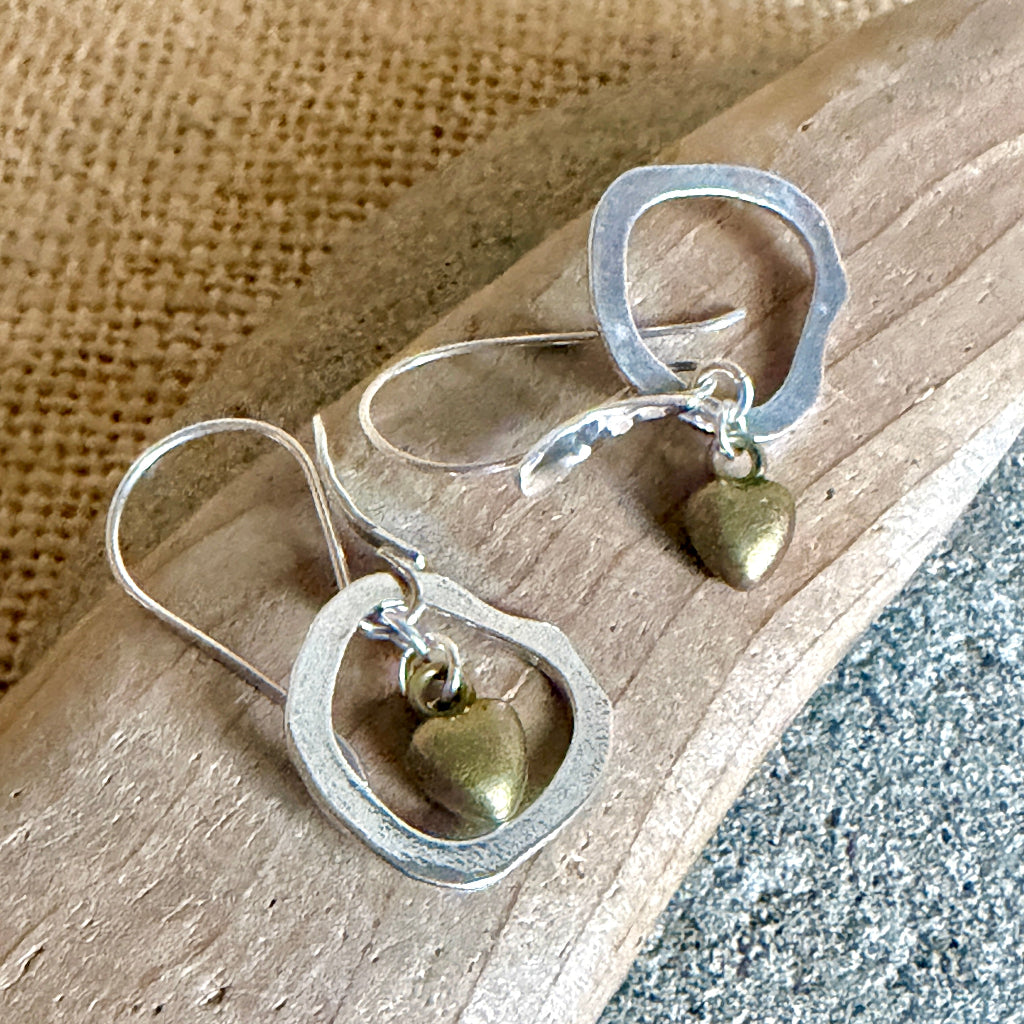 A pair of handmade dangle earrings with irregular silver-tone open circular frames and small antique bronze heart charms, displayed on a piece of driftwood over a burlap and stone background.