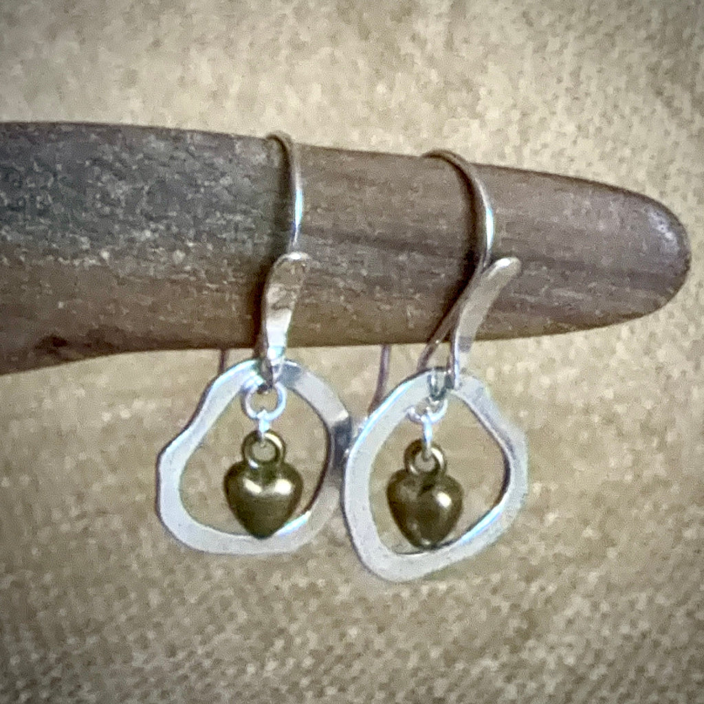 A pair of silver-toned dangle earrings with open teardrop-shaped hoops and small dangling heart charms, displayed on a curved wooden stick against a textured beige fabric background.