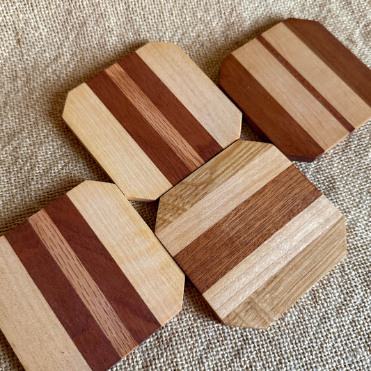 Four octagonal wooden coasters with striped patterns of light and dark wood grain, arranged on a textured beige burlap surface.