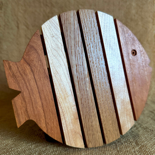 A fish-shaped wooden trivet made of vertical slats in two natural wood tones, photographed against a textured tan fabric background.