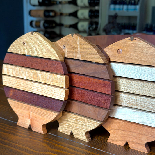 Three wooden fish trivets made from different wood species and tones, arranged on a dark wooden table with a blurred wine rack in the background.