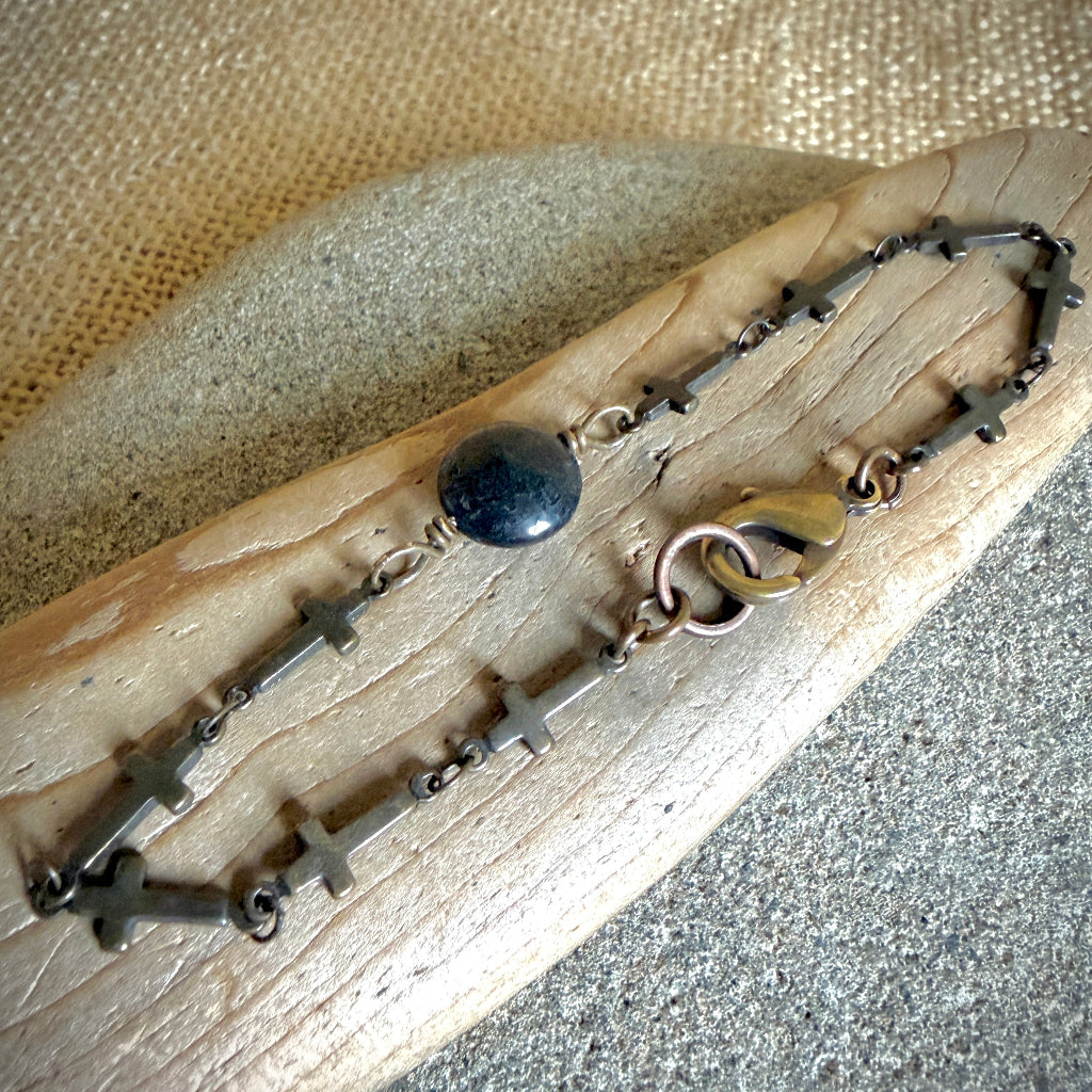A dark metal chain bracelet with multiple small cross charms, a round black shungite centerpiece, and a bronze-tone clasp, displayed on a curved piece of wood over a textured neutral background.