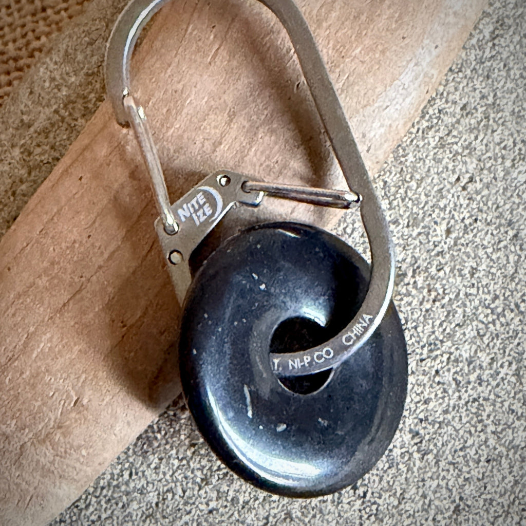 A close-up of a black shungite donut attached to a silver metal carabiner clip, resting on a piece of wood over a textured gray surface.
